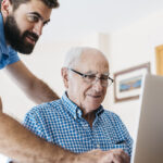Adult grandson teaching his grandfather to use laptop
